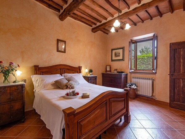 Un dormitorio con cama de madera, mesitas de noche, cómoda, ventana con vistas y techo con vigas a la vista.