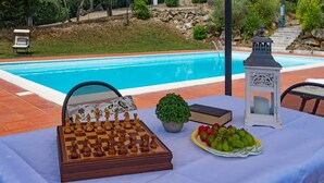 Escena junto a la piscina con un tablero de ajedrez, un libro, una planta en maceta y una cesta de frutas sobre una mesa.
