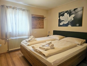 A double bed with neatly folded towels, a radiator, a wooden headboard, a wall-mounted light, and a painting of a flower.