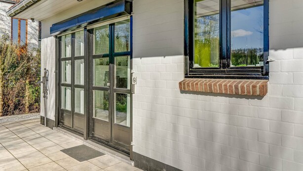 A building with a white brick exterior, large glass doors, and a small window with a view of greenery. - Holiday Home in Heuvelland With Garden (Heuvelland)
