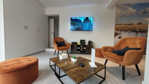 A modern living room with two orange armchairs, a wooden coffee table, and a large flat-screen TV mounted on the wall.
