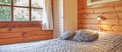 Habitación de cabaña de madera con una cama, una ventana con cortinas, un armario blanco y un cuadro en la pared.