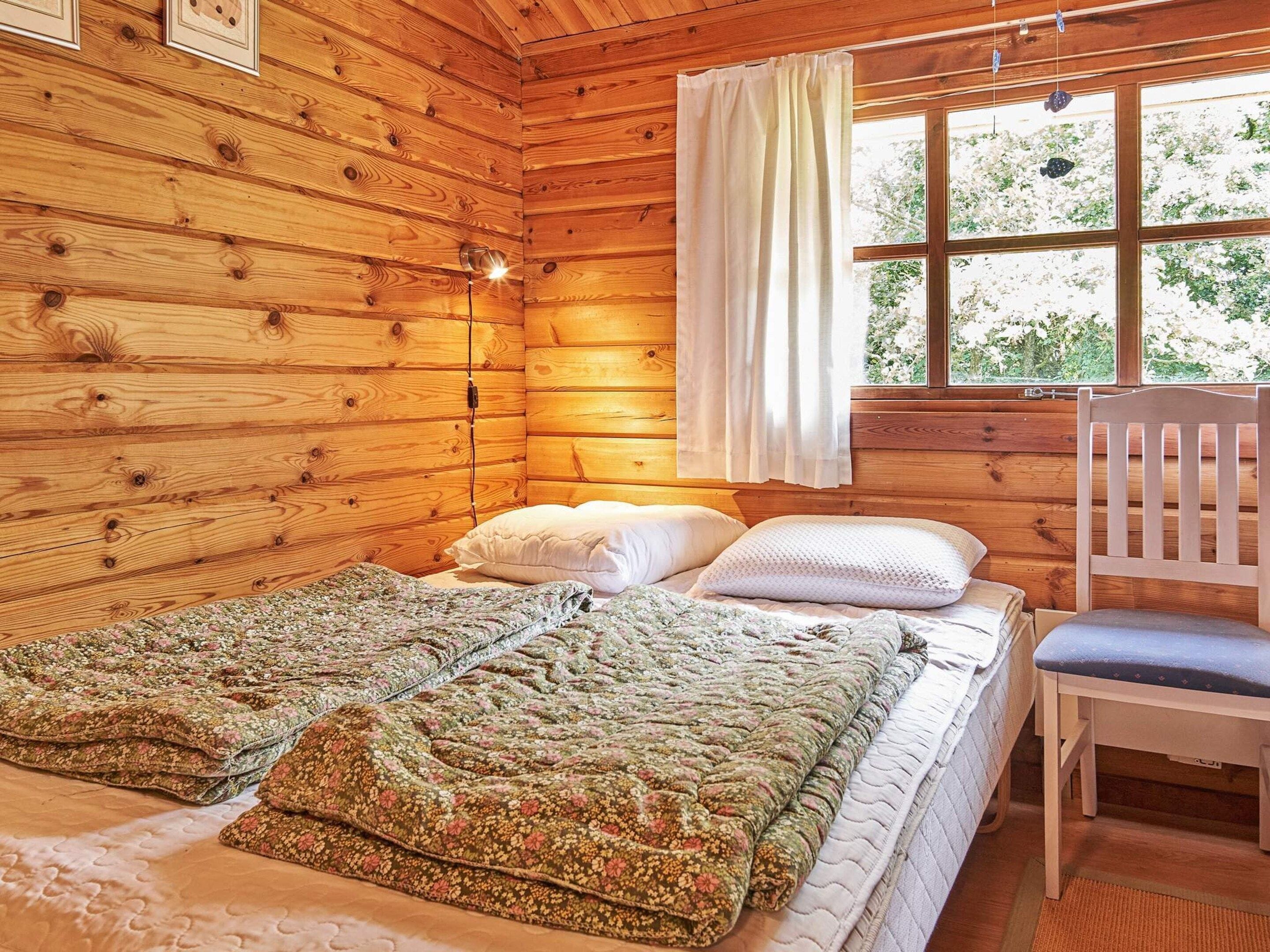A wooden cabin room with a bed, two pillows, a chair, and a window with curtains.