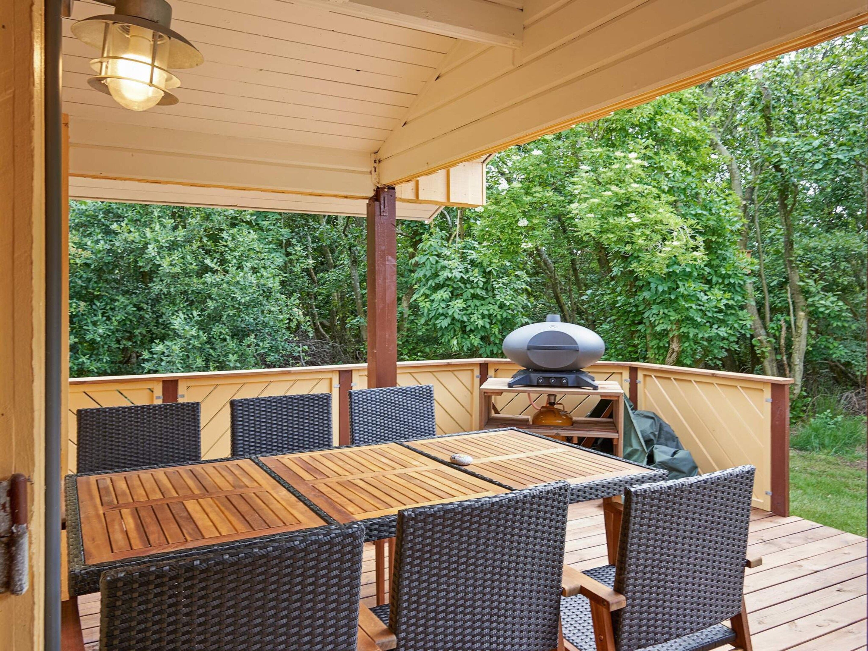 An outdoor patio with a wooden table and wicker chairs, a barbecue grill, and a view of a wooded area.