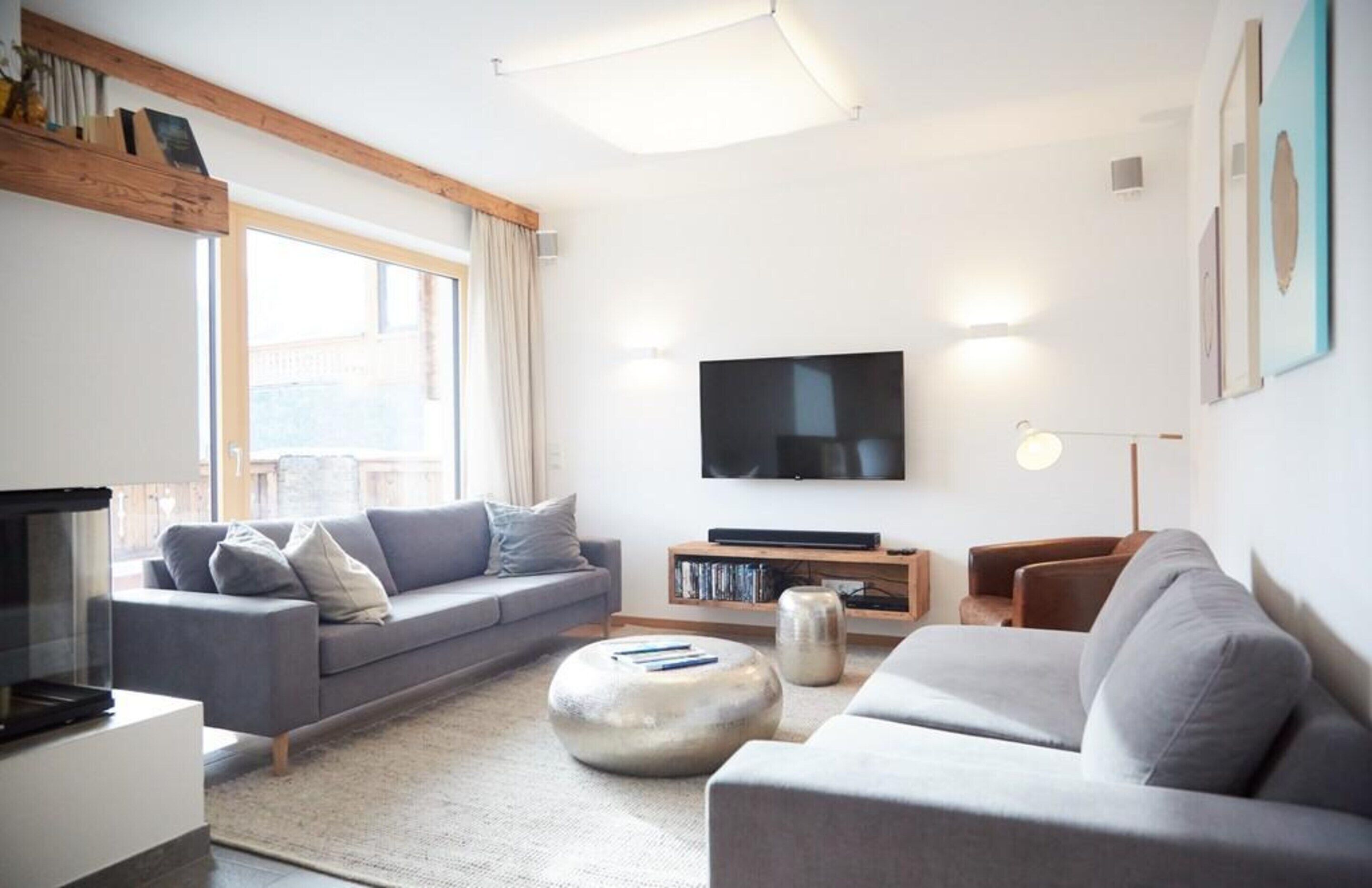 Ein modernes Wohnzimmer mit grauem Sofa, rundem Hocker, Flachbildfernseher und Bücherregal.