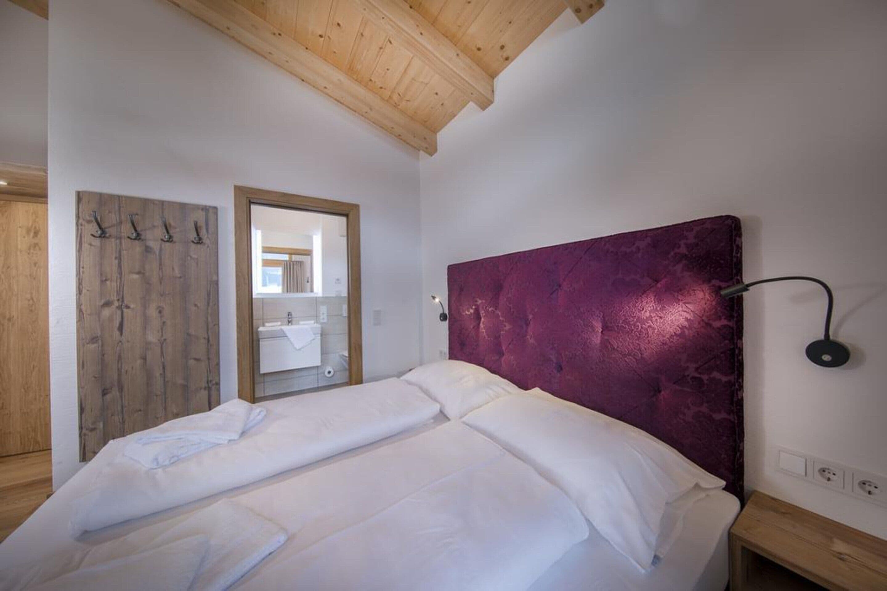 Une chambre avec un plafond en bois, un lit recouvert de draps blancs, une tête de lit violette et un miroir au mur.