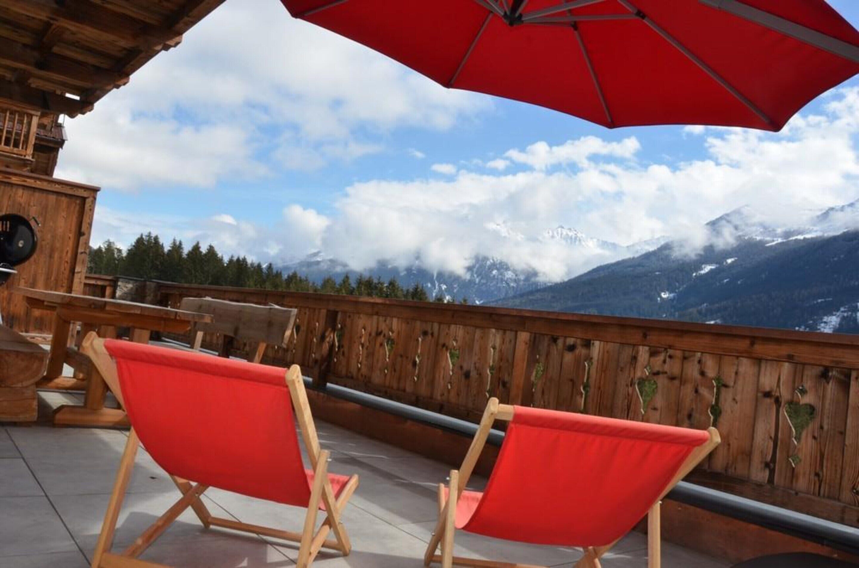A wooden deck with red chairs and a red umbrella, overlooking a mountainous landscape.