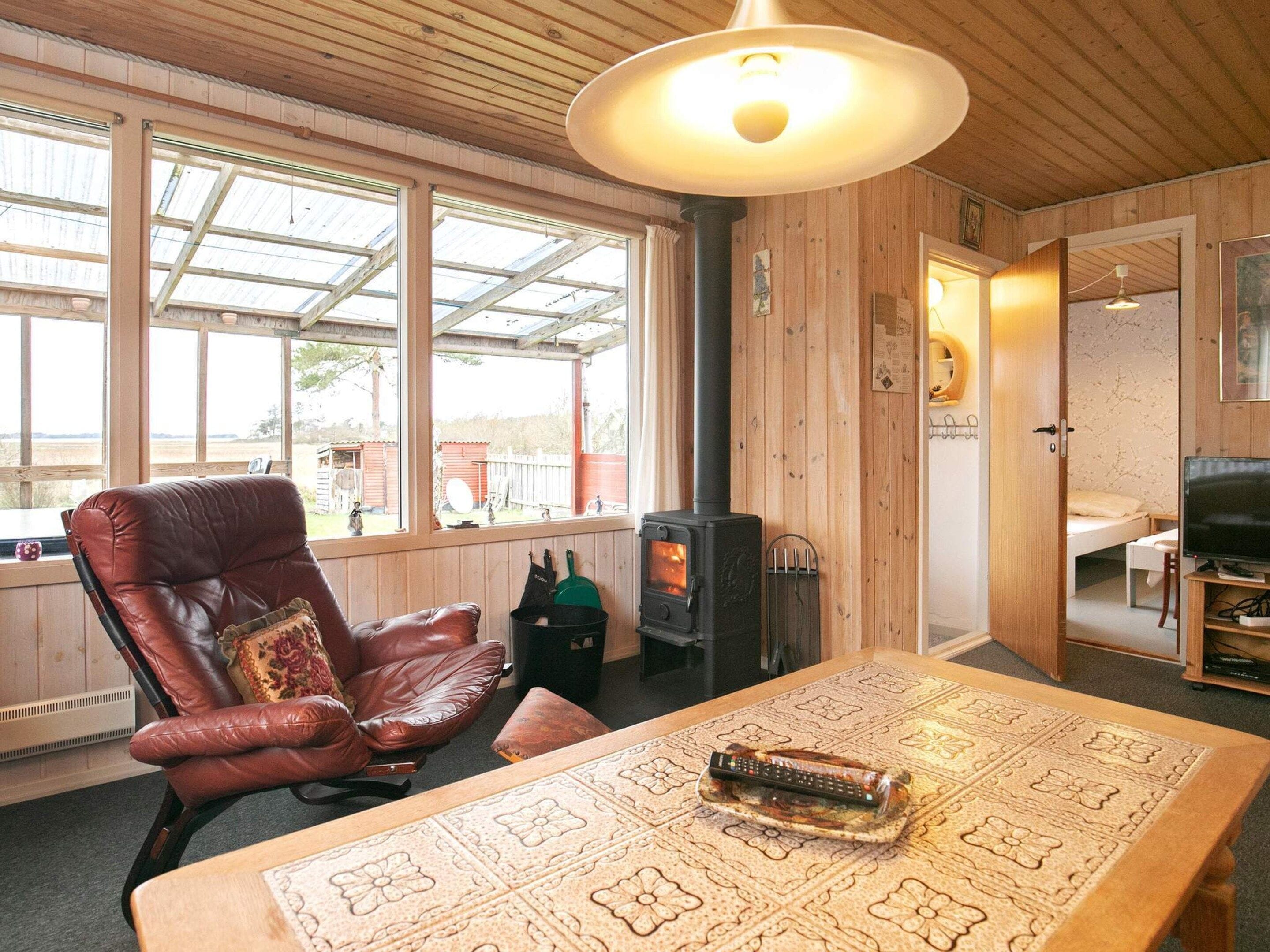 A cozy room with a wooden ceiling, a large window, a leather chair, a table with a remote control, and a television.