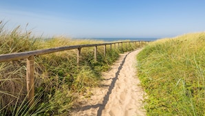 Un sendero arenoso con barandillas de madera, rodeado de hierba y que conduce a una playa.