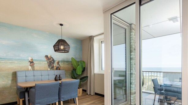 Un comedor con una mesa de madera y sillas tapizadas en azul, una planta grande en maceta y un mural en la pared con una escena de playa.