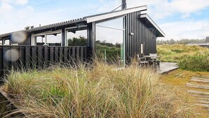 A modern, minimalist building with large windows and a flat roof, situated in a natural landscape with tall grasses. - Holiday Home in Saltum (Saltum)