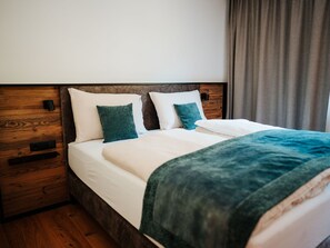 Un lit bien fait, avec du linge de lit blanc et un plaid turquoise, une tête de lit en bois et une table de chevet en bois sur laquelle se trouve un objet noir.