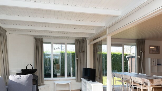 A modern living room with a grey sofa, a TV on a white stand, and a dining area with a table and chairs. - Attractive House with Sunny Garden near Veerse Meer (Kortgene)