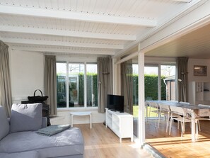A modern living room with a grey sofa, a TV on a white stand, and a dining area with a table and chairs. - Attractive House with Sunny Garden near Veerse Meer (Kortgene)