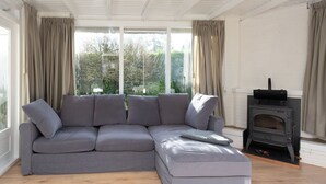 A modern living room with a grey sectional sofa, large windows with curtains, and a wood-burning stove. - Attractive House with Sunny Garden near Veerse Meer (Kortgene)