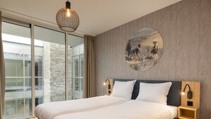 Ein modernes Schlafzimmer mit einem großen Bett, einem runden Wandkunstwerk und Blick auf ein Backsteingebäude durch große Fenster.