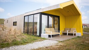 A modern house with a wooden exterior, large glass windows, and a yellow section. There are two outdoor seating areas with chairs. - Lodge in Callantsoog Near North Sea Beach (Callantsoog)