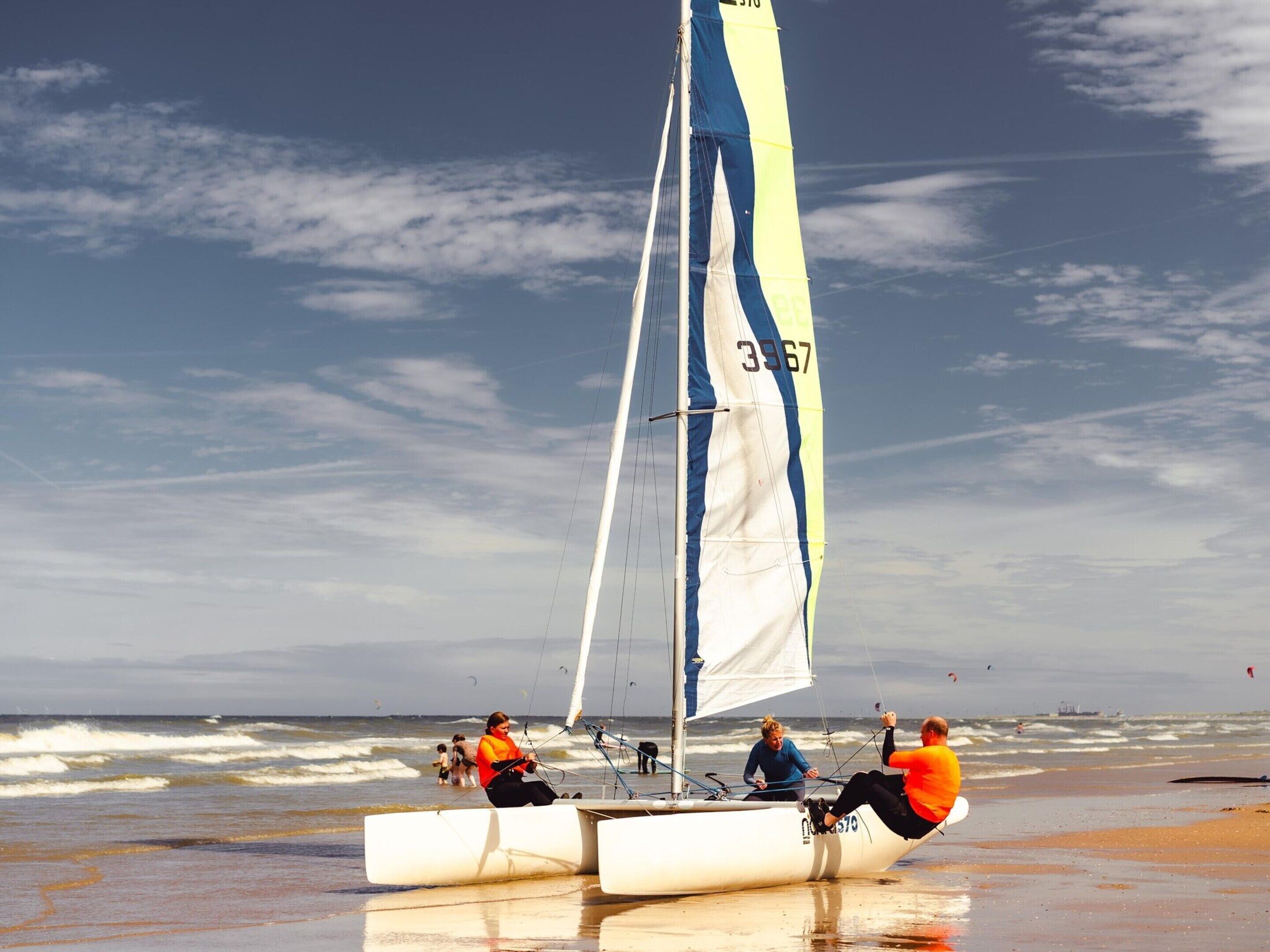 A sailboat with two people on the beach, a sandy shore, and the ocean in the background.