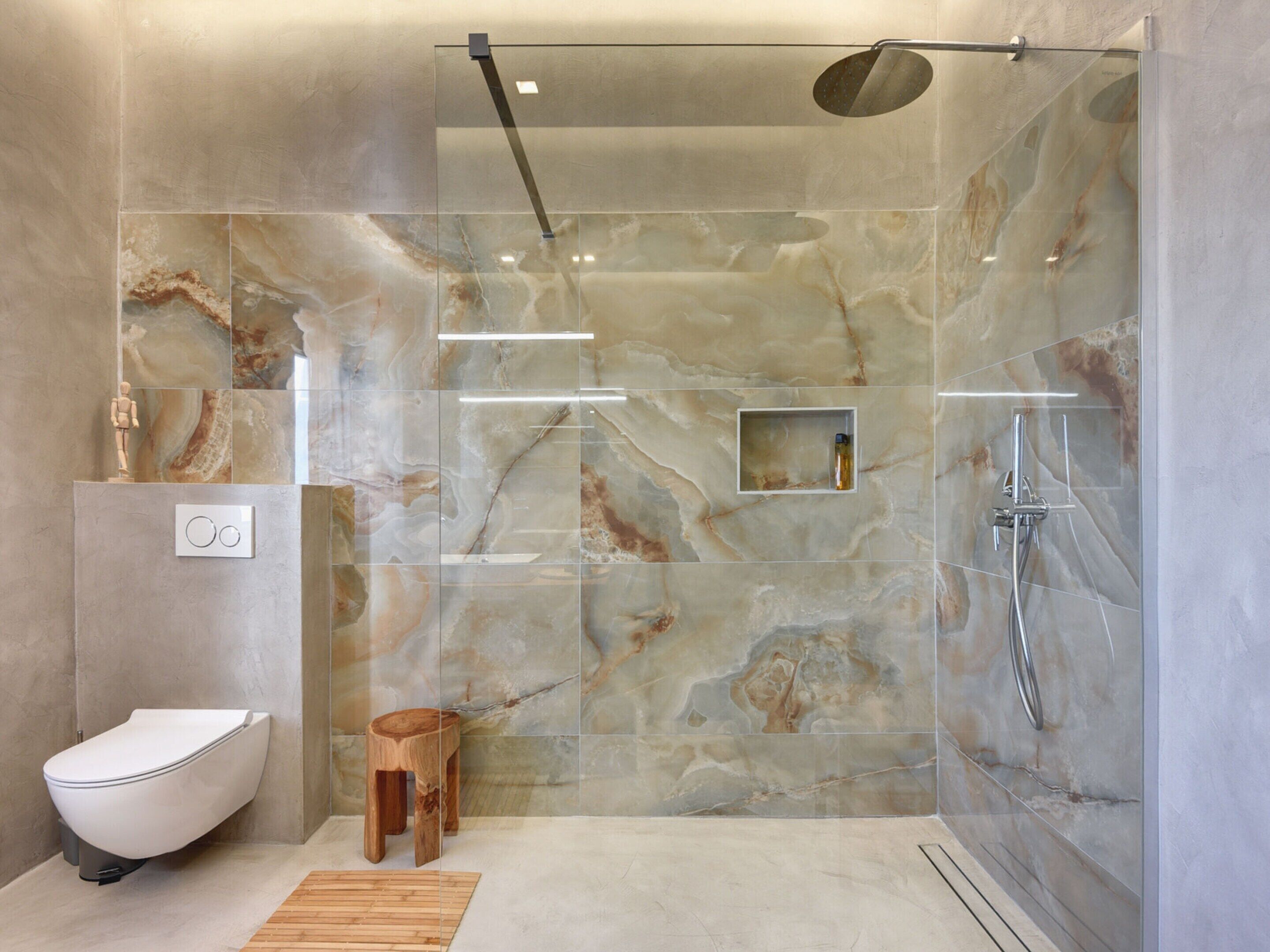 A modern bathroom with a marble shower wall, a freestanding toilet, and a wooden stool.
