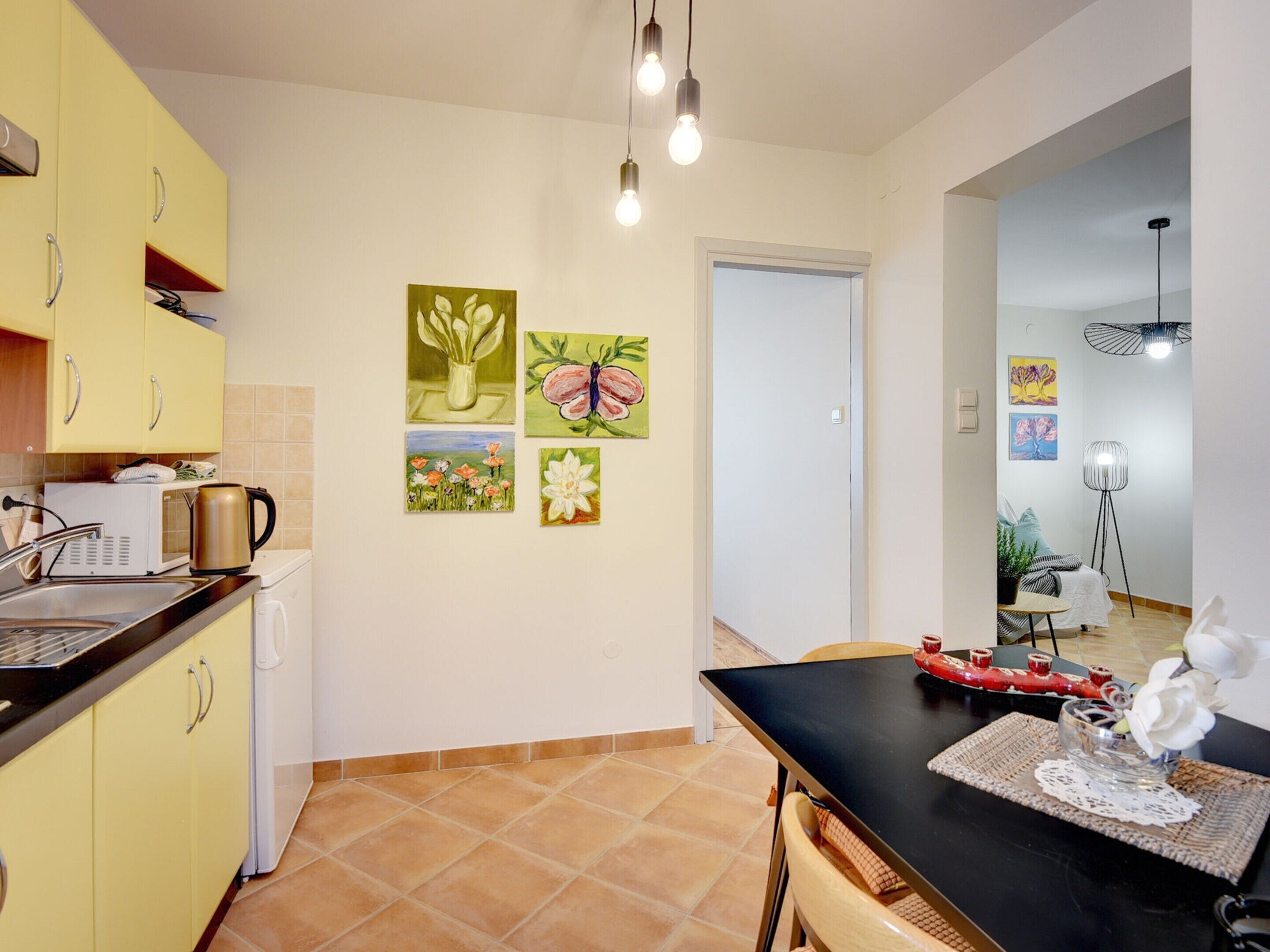 A compact kitchen with yellow cabinets, a black countertop, and a dining area with a table and chairs. There are four wall-mounted paintings depicting flowers and a butterfly.