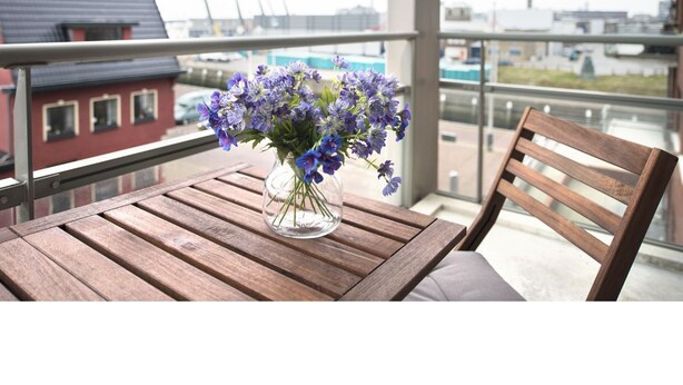 A balcony with a wooden table and chairs, a vase of purple flowers, and a view of buildings and a bridge in the background.