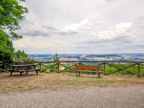 Un área de picnic con banco y mesa, rodeada por una valla de madera, con vistas a un paisaje pintoresco con colinas y campos.