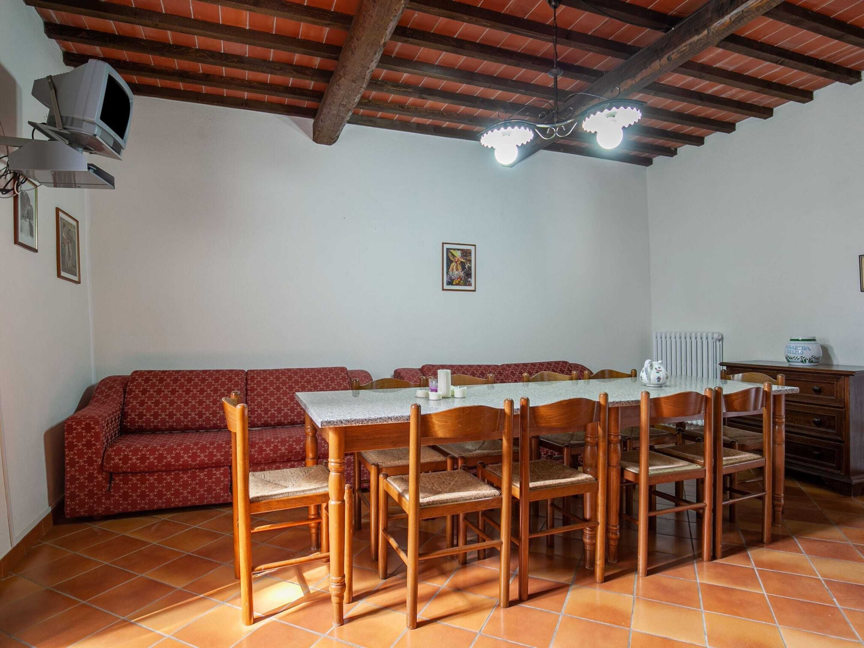 Ein Essbereich mit einem langen Holztisch, sechs Stühlen, einem roten, gemusterten Sofa und einer Decke mit freiliegenden Balken und Lampen.