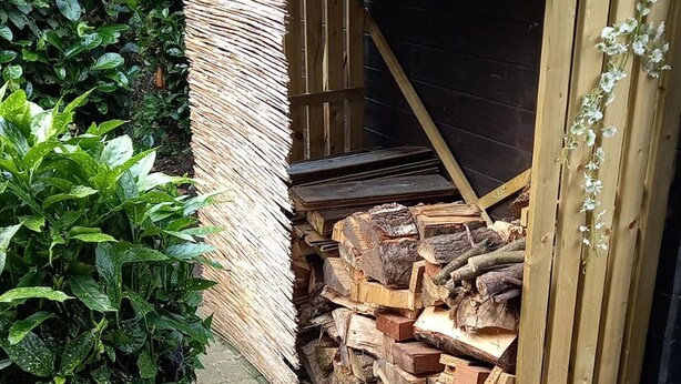 Una estructura de madera con un muro tejido y una pila de leña.