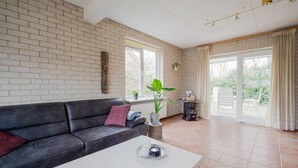 Una sala de estar con un sofá de cuero negro, una mesa de centro blanca y una pared de ladrillo. Hay ventanas con cortinas, una planta en el alféizar y vistas al exterior.