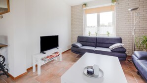 Una sala de estar con televisor de pantalla plana, un sofá gris, una mesa de centro blanca y una ventana con vistas al exterior.