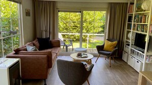 A spacious living room with a brown sofa, a grey armchair, a small round table, and a bookshelf filled with books. - Comfy Chalet with Palletstove near Wadden Sea (Hippolytushoef)