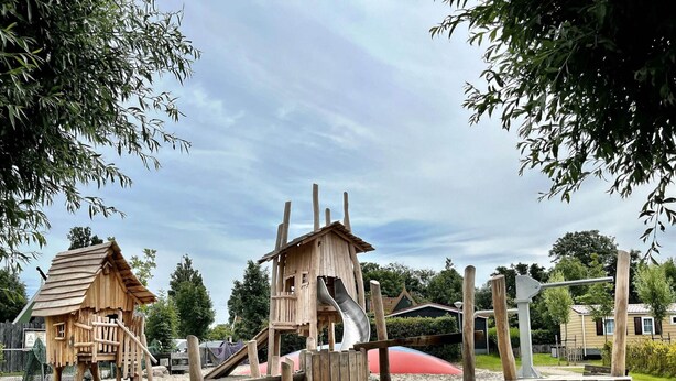 A playground with wooden structures, including a slide and climbing frames, surrounded by sand and trees. - Comfy Chalet with Palletstove near Wadden Sea (Hippolytushoef)
