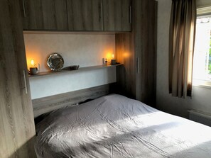 A bedroom with a bed, a shelf with decorative items, and a window with curtains. - Comfy Chalet with Palletstove near Wadden Sea (Hippolytushoef)