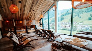 Une pièce avec des poutres en bois au plafond, de grandes fenêtres donnant sur un village et plusieurs fauteuils inclinables.