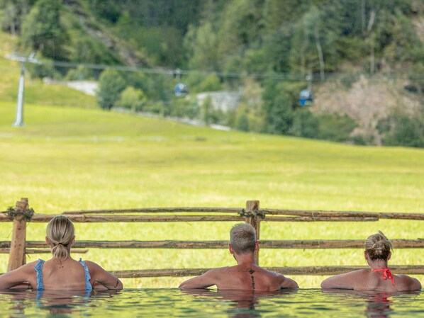 Drie personen in een natuurzwembad, met een houten omheining en groen in de achtergrond.