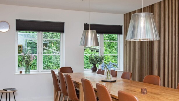 A dining area with a wooden table and brown chairs, large windows with black blinds, and a hanging pendant light. - Holiday Home in VÃ¦ggerlÃ¸se (Vaeggerlose)