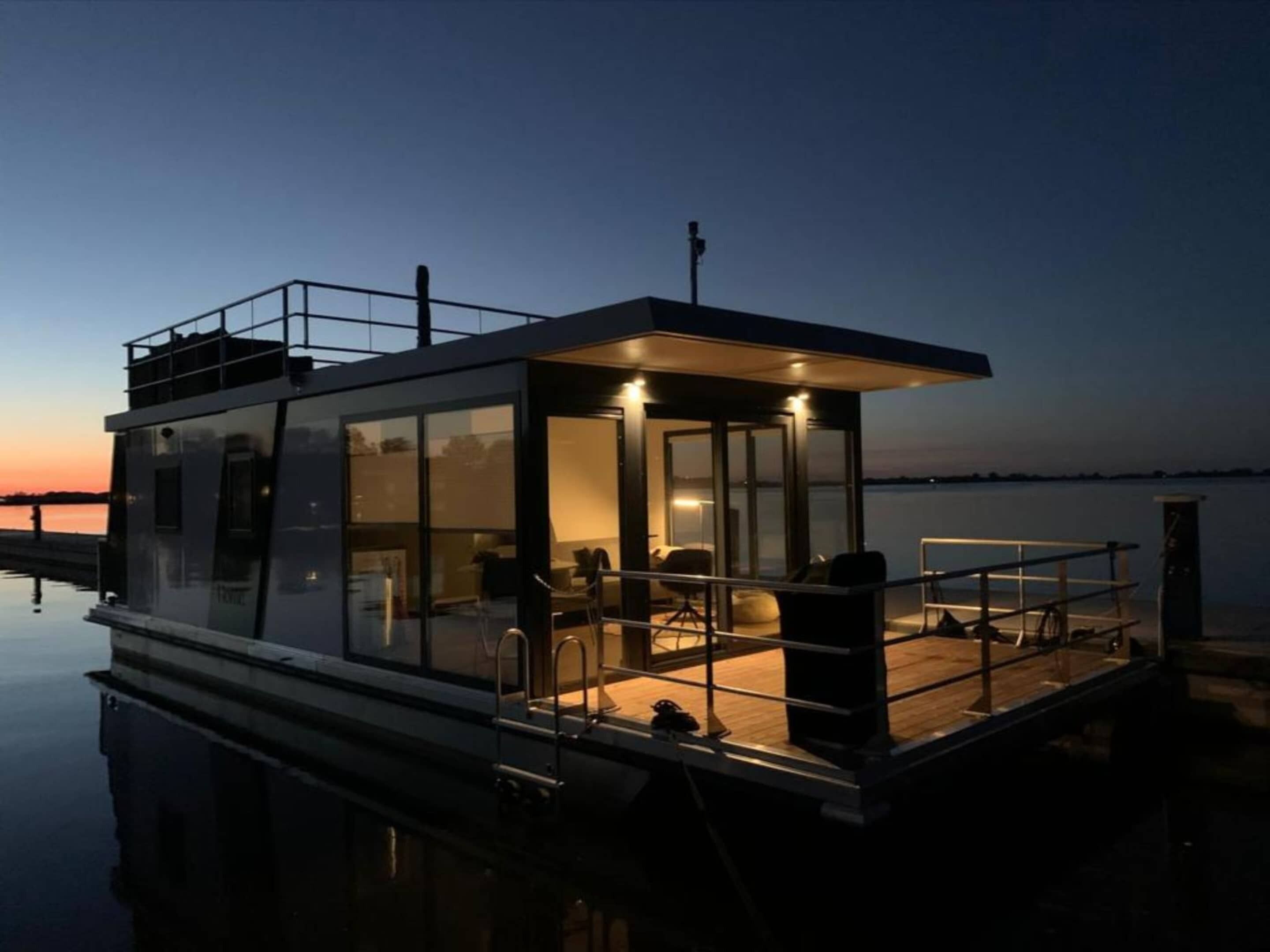 A modern floating house with a glass exterior, a covered deck, and a view of the water.