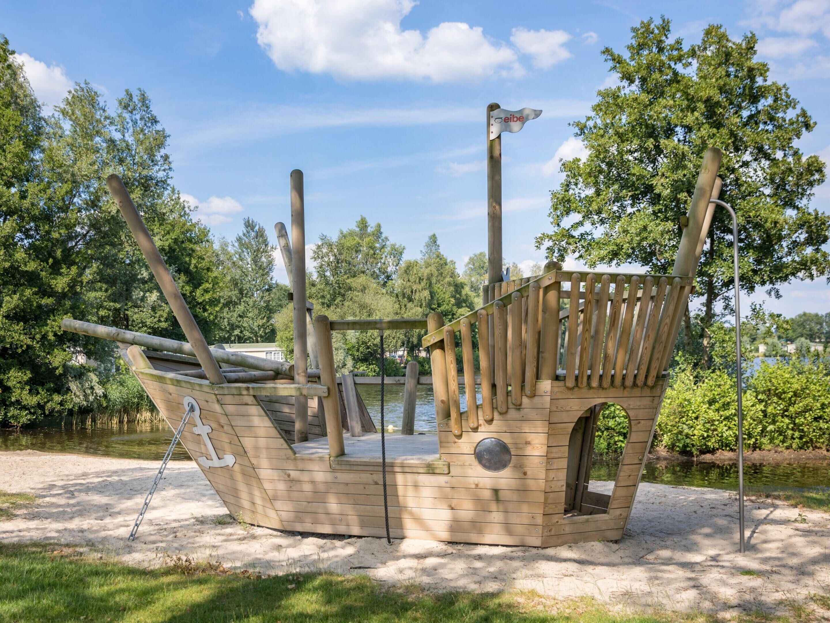 Une structure de jeu en forme de bateau de pirate en bois, dotée d’un toboggan, entourée d’arbres et bordée par un plan d’eau.