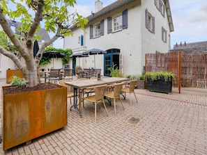 An outdoor seating area with a table and chairs, a tree planter, and a building with a white facade and shutters. - Garden Apartment in Grubbenvorst (Grubbenvorst)