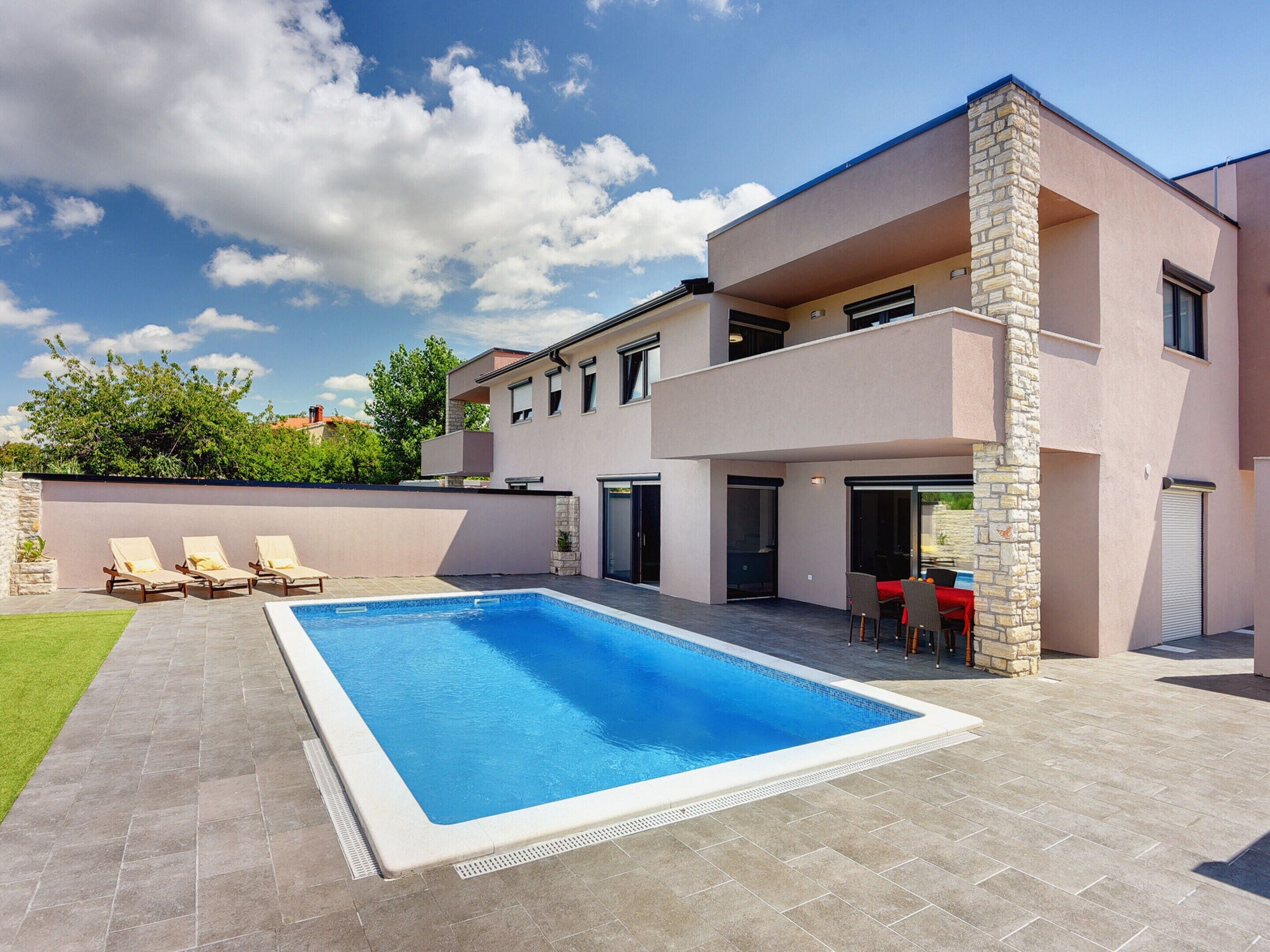Una casa moderna de dos pisos con piscina, mobiliario exterior y un pilar de piedra.