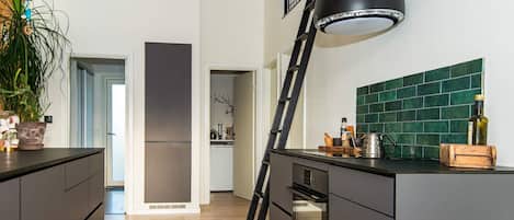 Una cocina moderna con armarios oscuros, una lámpara colgante negra, un salpicadero de azulejos verdes y una escalera de madera apoyada en la encimera.