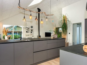 A modern kitchen with a central island, pendant lighting, and a view of a green house. - Holiday Home in Tarm (Tarm)