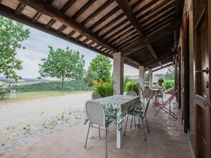 Um pátio externo com mesa e cadeiras, uma área coberta com teto de madeira, e vista para o campo.