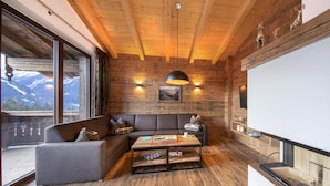 Een gezellige kamer met een houten plafond, een grijze bank, een houten bijzettafeltje en een groot raam met uitzicht op de bergen.