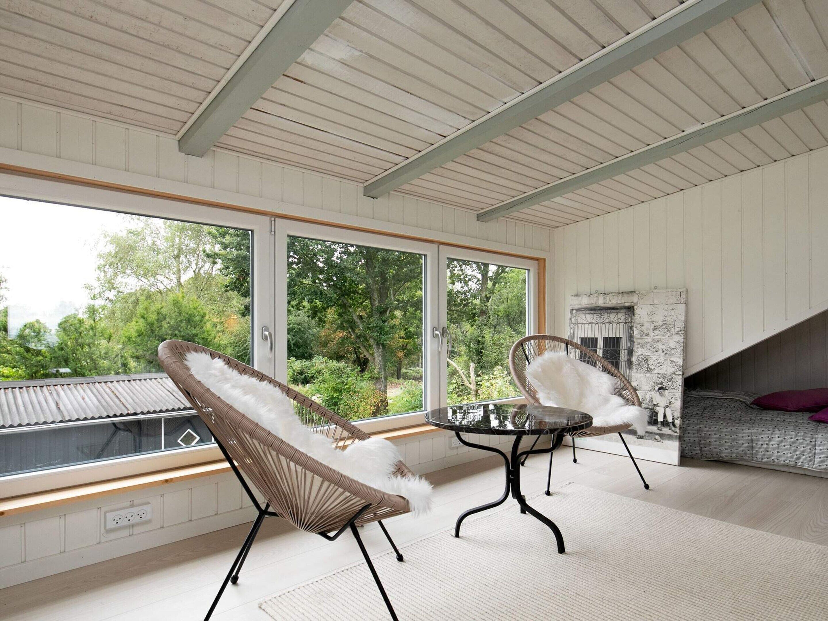 Ein Zimmer mit einem großen Fenster, zwei Korbstühlen, einem kleinen runden Tisch und einer Couch mit einem gemusterten Kissen.