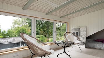 Ein Zimmer mit einem großen Fenster, zwei Korbstühlen, einem kleinen runden Tisch und einer Couch mit einem gemusterten Kissen.