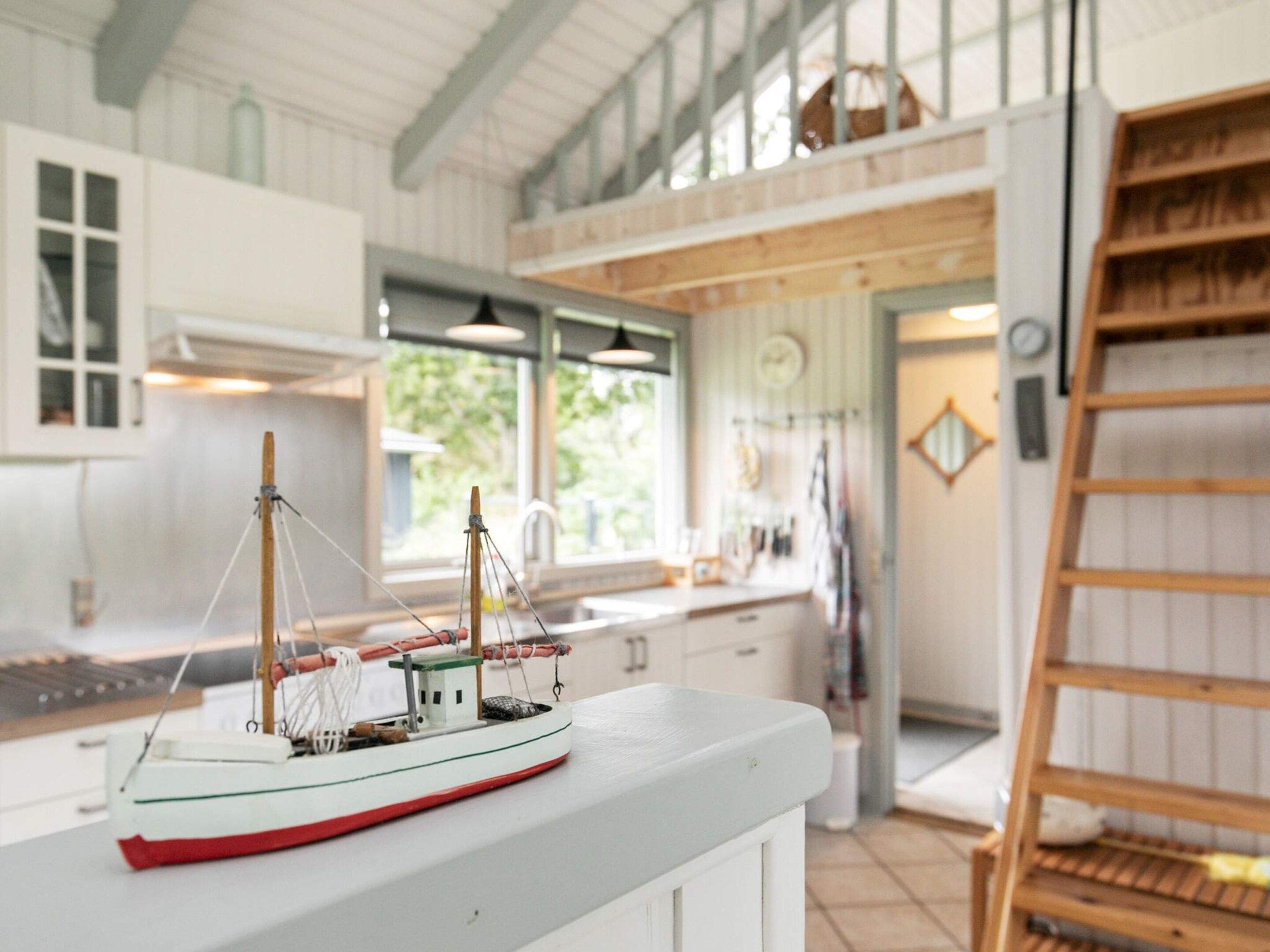 A model ship on a counter in a well-lit kitchen with large windows, a wooden ladder, and a white interior.