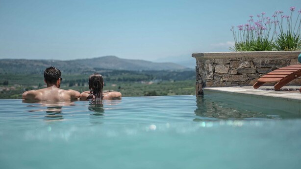 A couple relaxing in a private infinity pool with a stone wall and a wooden lounge chair, overlooking a mountainous landscape.