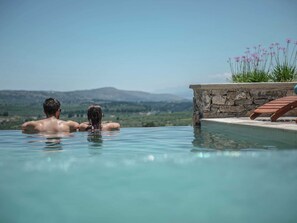 Un couple se détend dans une piscine à débordement privée, bordée d’un mur de pierre et agrémentée d’une chaise longue en bois, avec une vue imprenable sur un paysage montagneux.