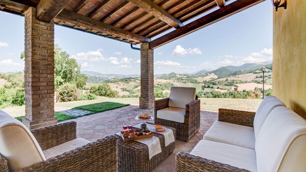 A covered outdoor seating area with wicker furniture and a view of rolling hills. - Holiday Home in Urbania With Hill Views (Urbania)